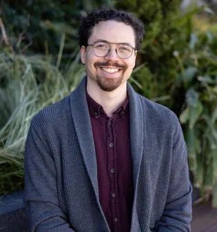 Closeup photograph of Tyler, taken by Matthew Washburn, smiling, wearing a gray cardigan over a maroon button up shirt, sitting on a bench in front of some foliage.